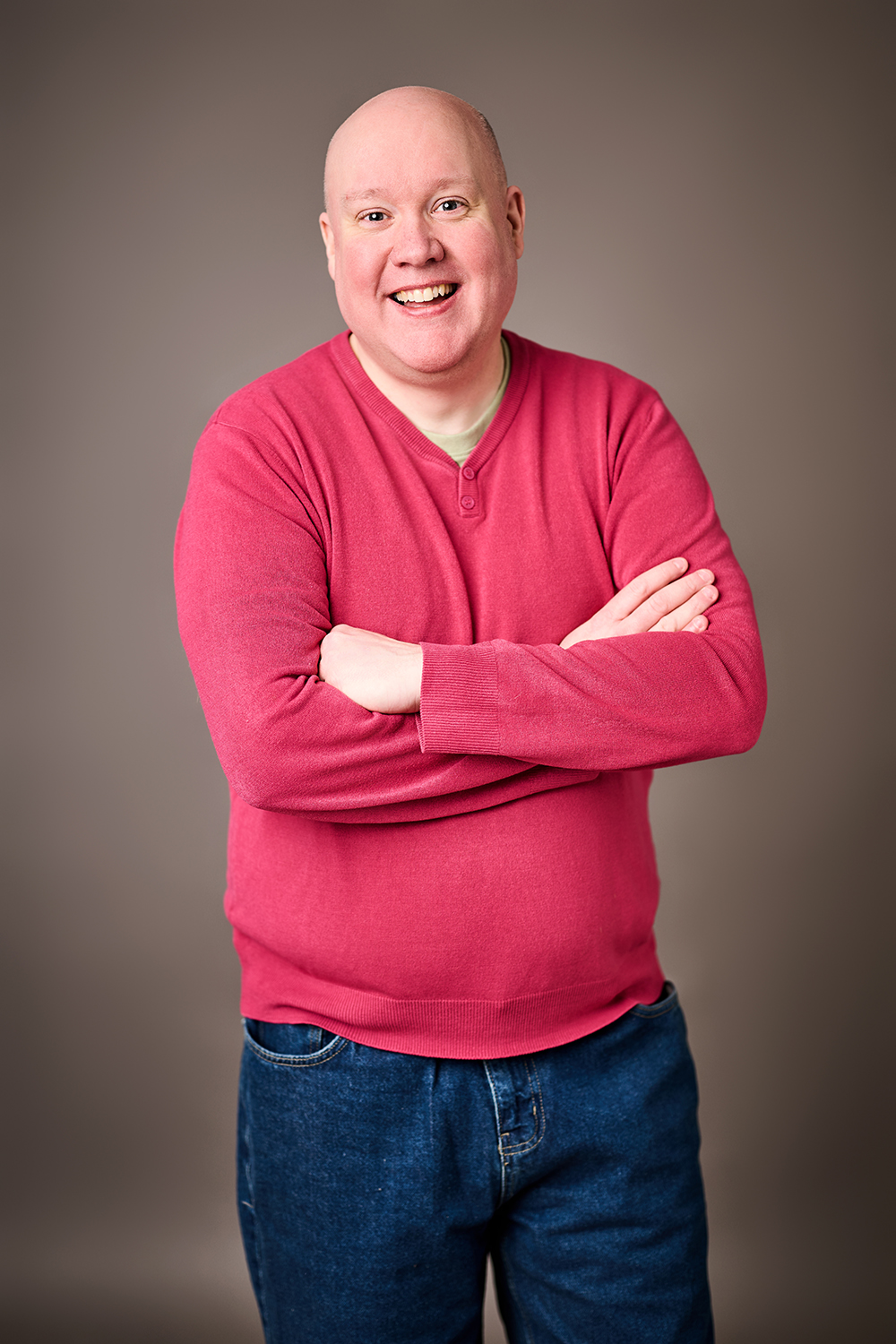 A bald white man in a pink jumper smiles towards the camera, arms folded.